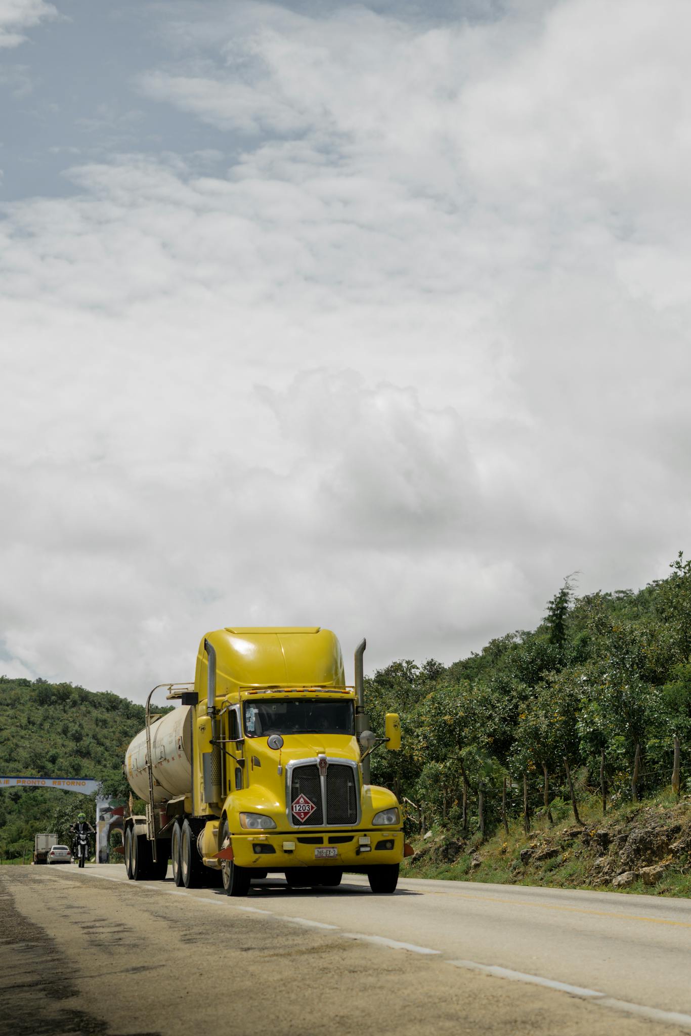 A yellow freight truck travels on a rural road under a cloudy sky.